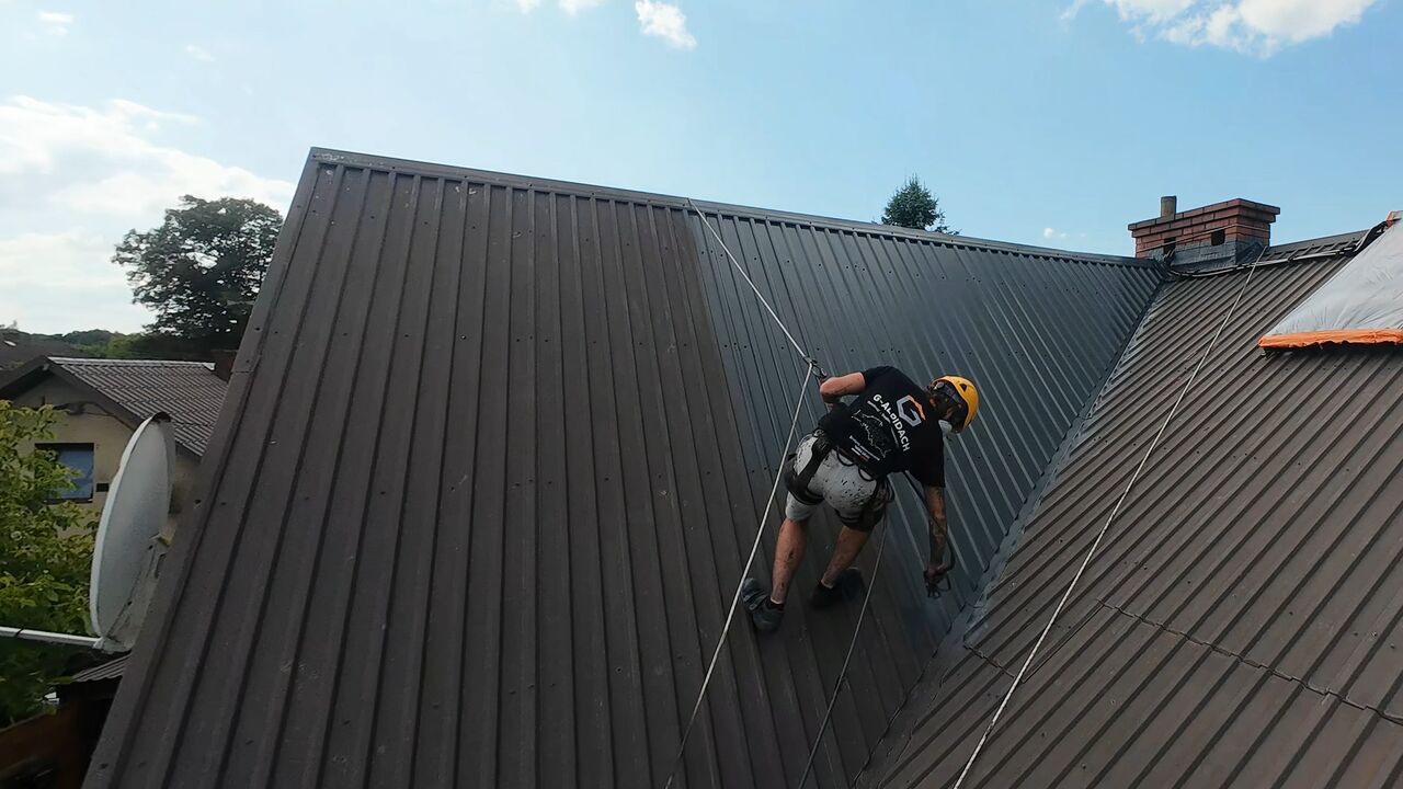 Malowanie dachu z blachy — pracownik G-ALPIDACH w uprzęży pracuje na ciemnym dachu z blachy trapezowej